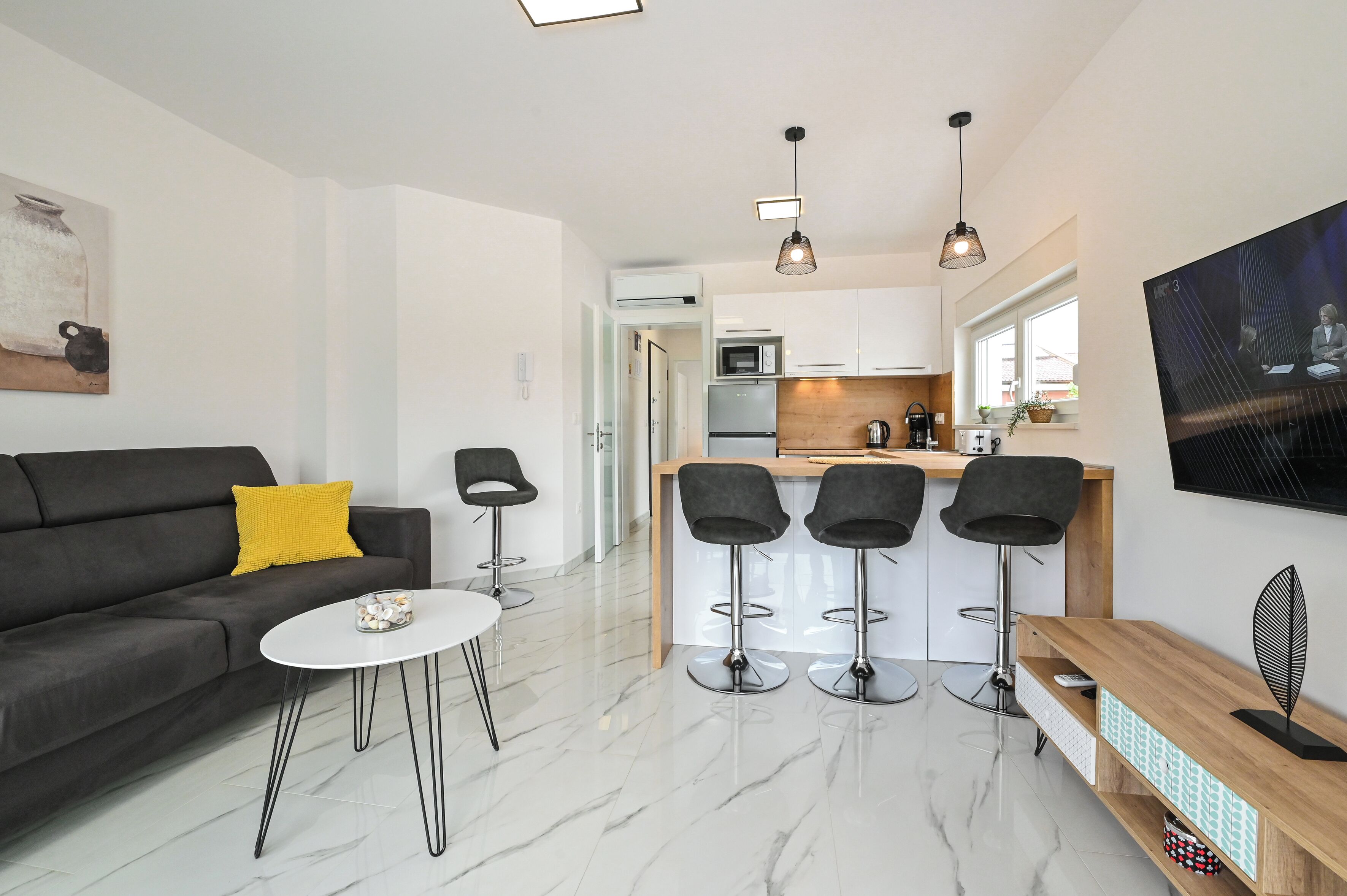 A modern living room with a sofa, coffee table, and bar stools. It includes a kitchen area with a microwave, refrigerator, and bar counter.