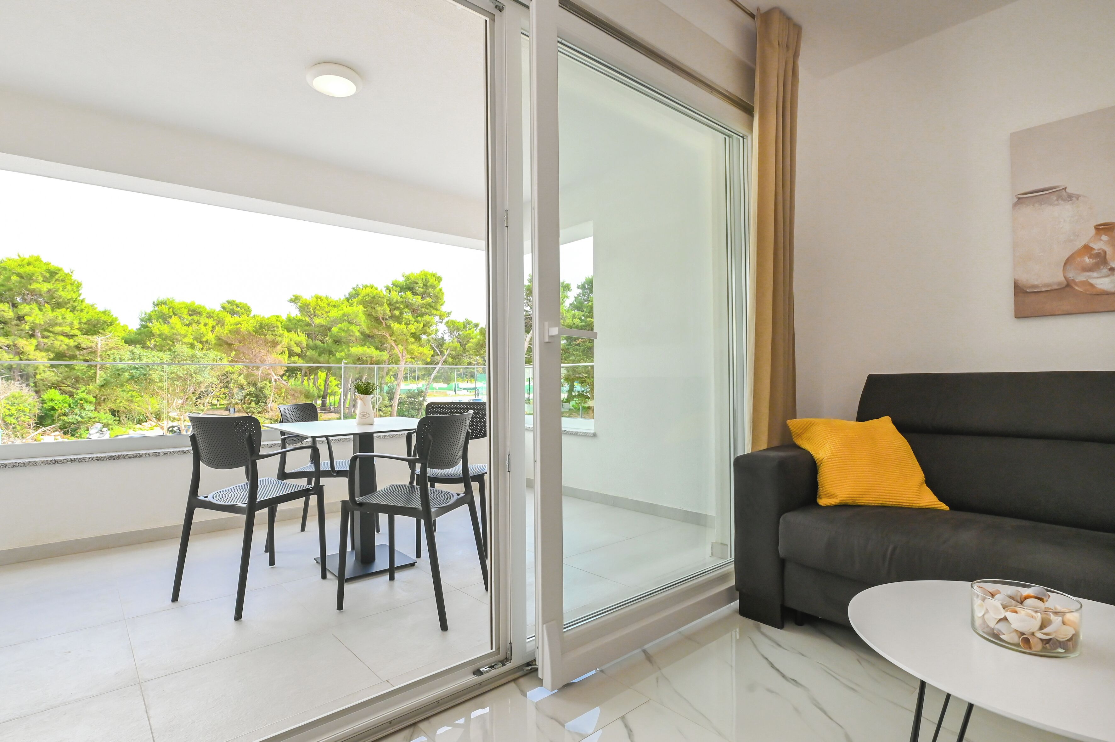 Una sala de estar moderna con un ventanal que da a un balcón, un sofá negro con un cojín amarillo y una mesa de vidrio con un cuenco de conchas.