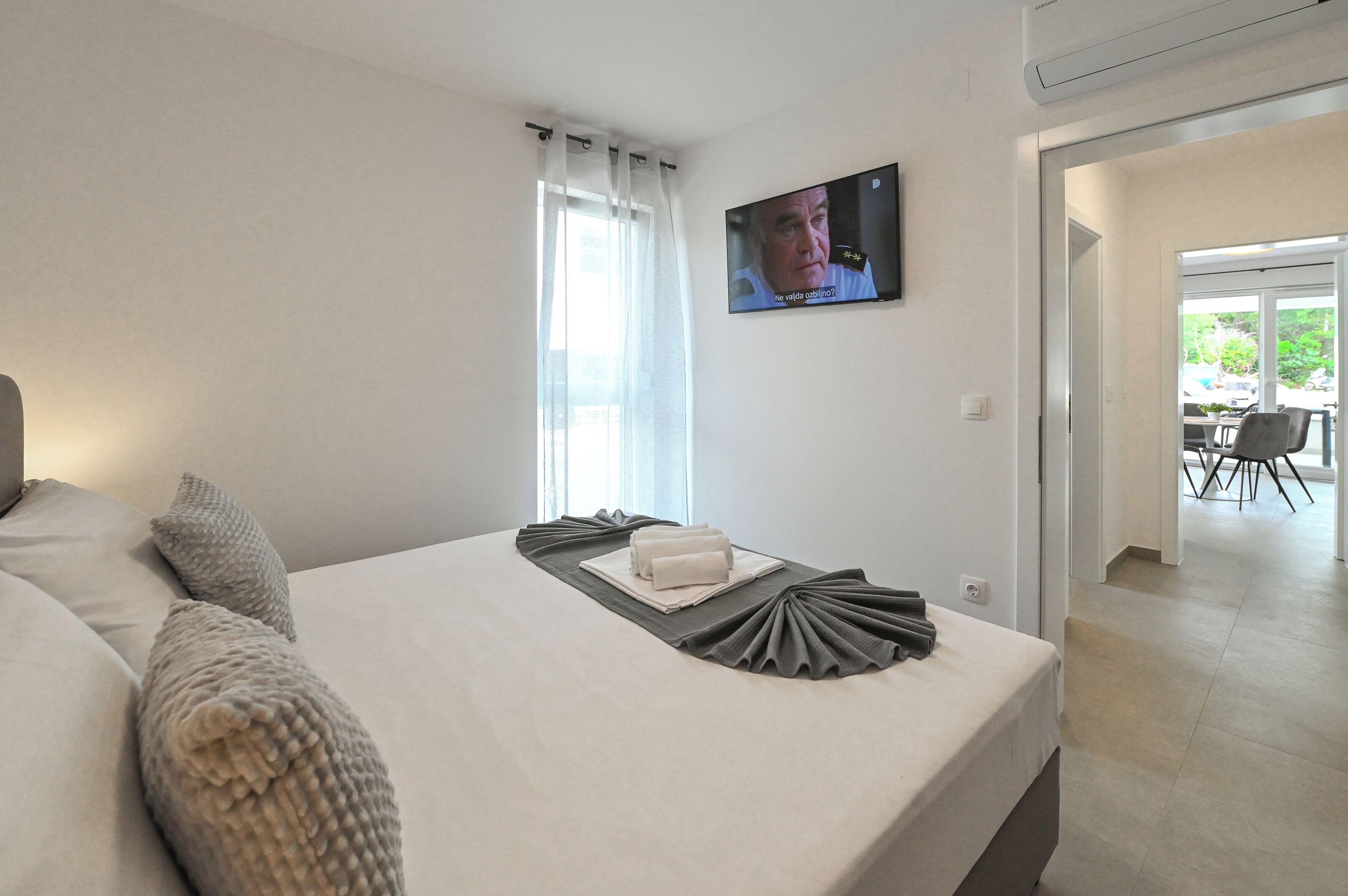 A modern hotel room with a neatly made bed, a flat-screen TV mounted on the wall, and a view of the outdoor area through the window.