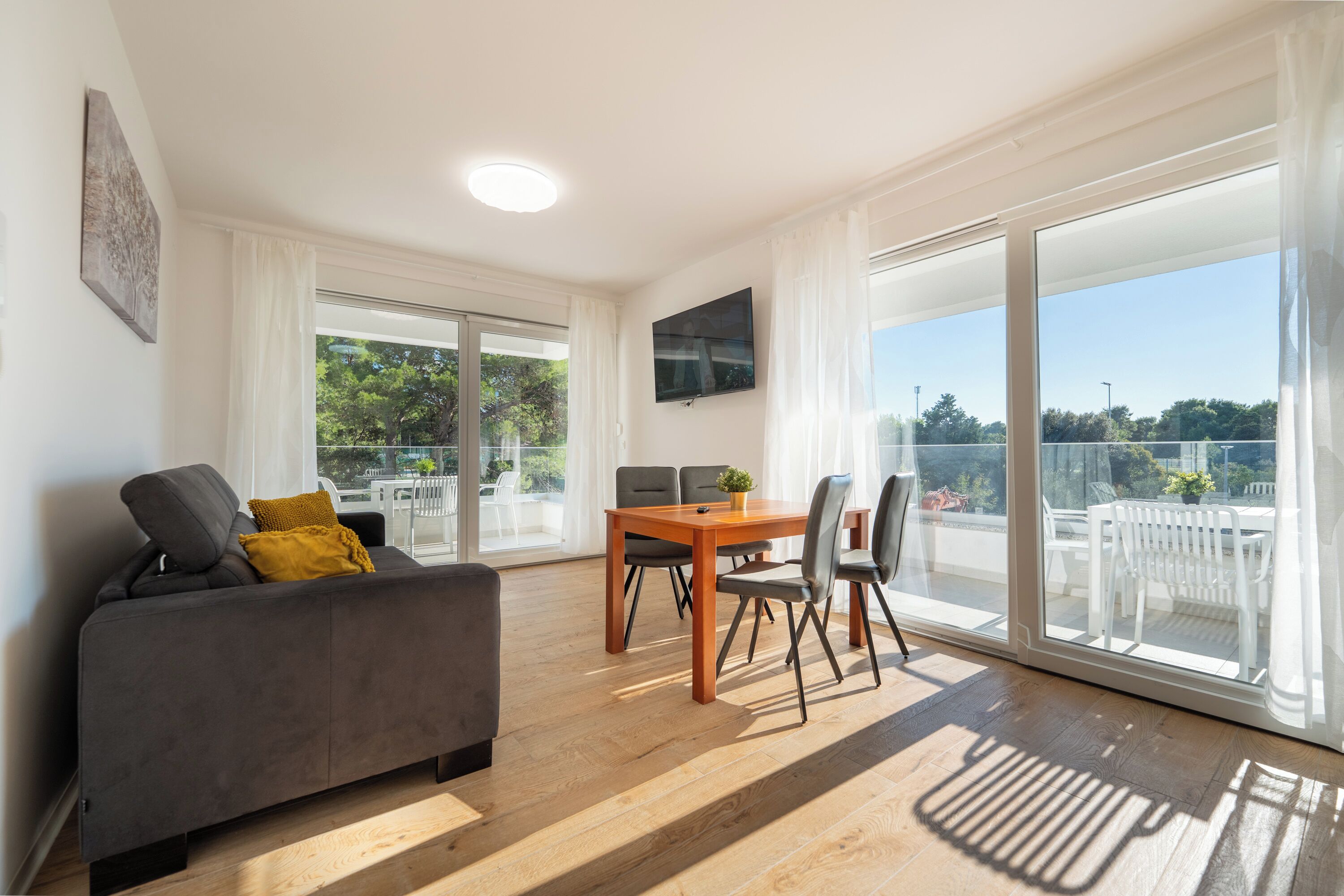 Ein modernes Wohnzimmer mit einer Couch, einem Esstisch und Stühlen. Ein Fernseher ist an der Wand befestigt und es gibt große Fenster mit Vorhängen.