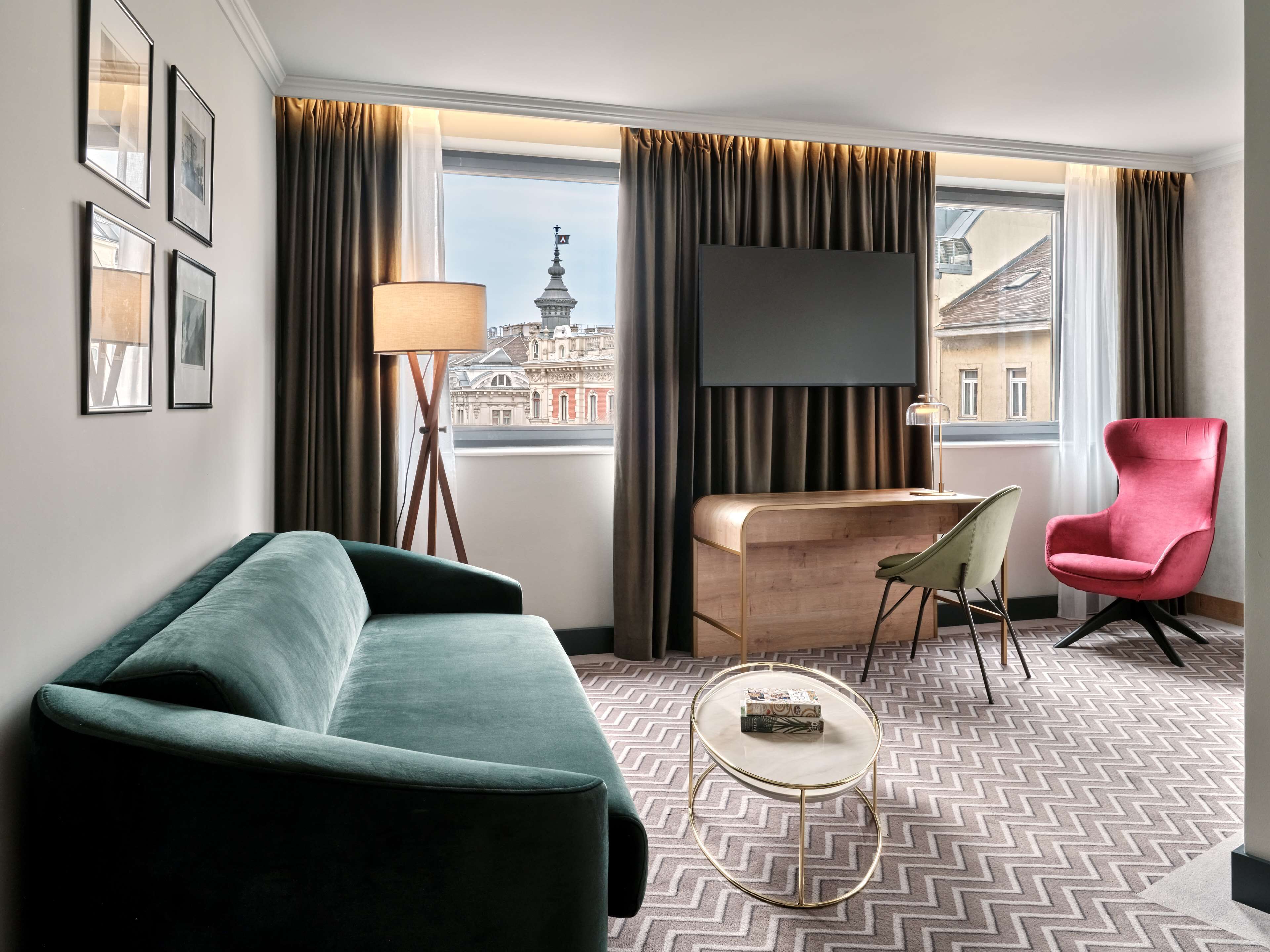 Ein Hotelzimmer mit einem grünen Sofa, einem roten Sessel, einem kleinen runden Tisch, einem Flachbildfernseher und Blick auf ein Gebäude mit einem Turm.