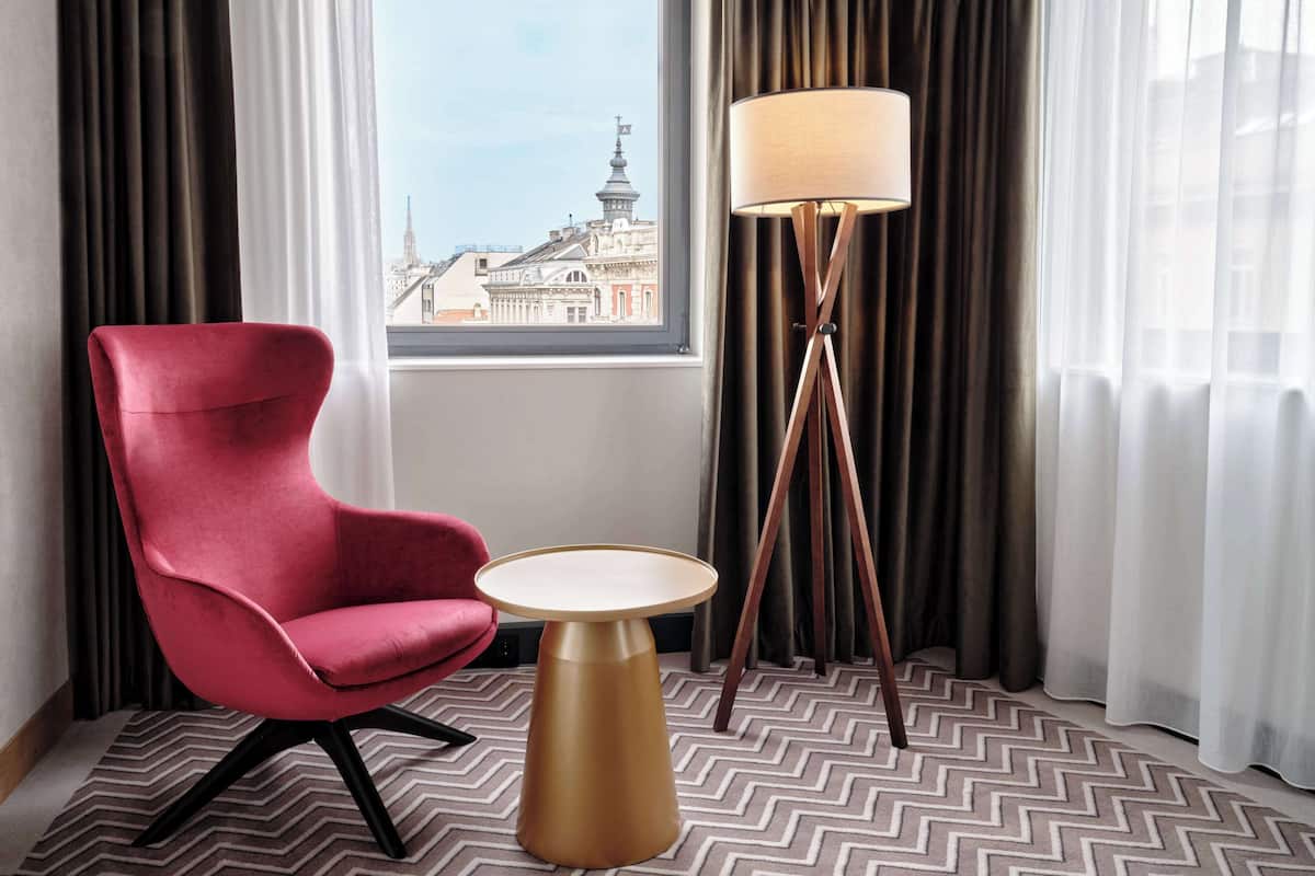 Ein Hotelzimmer mit einem roten Sessel, einem kleinen runden Tisch, einer Stehlampe und Blick auf eine Stadtansicht durch ein Fenster.