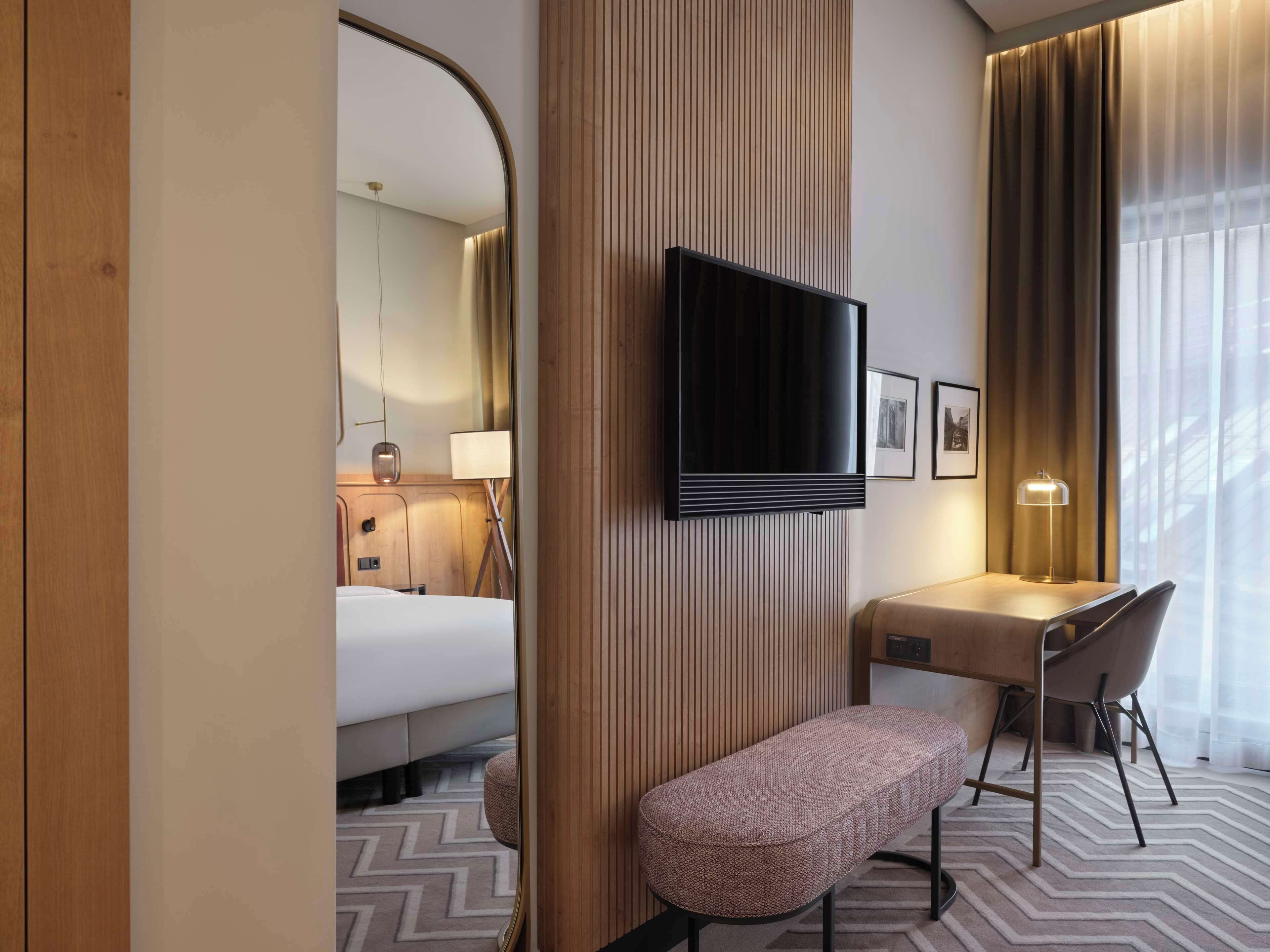 Habitación de hotel con una cama, un escritorio con silla, un banco, un televisor en la pared y una ventana con cortinas.