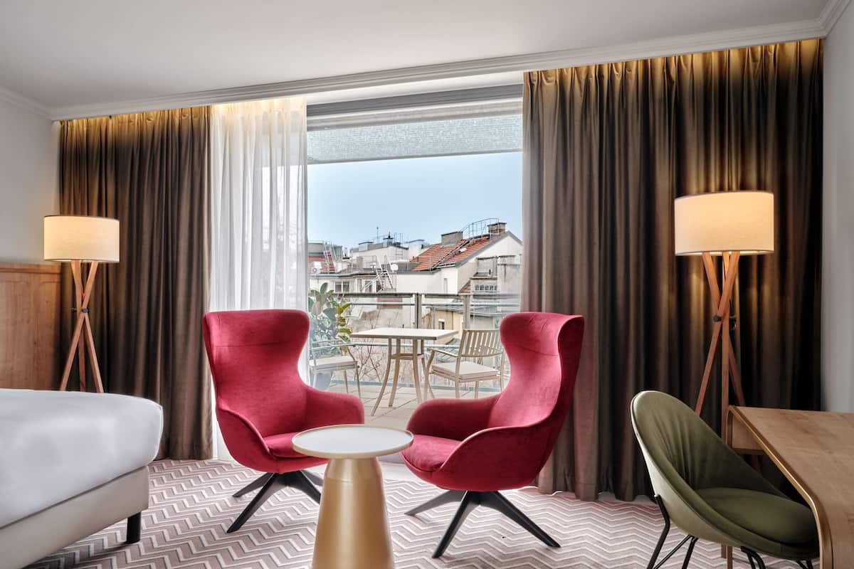 Ein Hotelzimmer mit einem Bett, zwei roten Sessel, einem kleinen runden Tisch, einem grünen Stuhl und zwei Stehlampen.