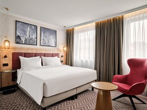 Una habitación de hotel con una cama grande, un sillón rojo, una mesita redonda, dos lámparas de pared y dos cuadros en la pared.