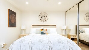 Quarto de hotel com cama, duas mesas de cabeceira, um espelho grande e um objeto decorativo na parede.