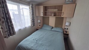 Un dormitorio con cama, mesita de noche, estante en la pared con un cartel y ventana con cortinas.