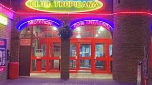 Un edificio con un letrero de neón que dice "CLUB TROPICA" y dos entradas, una indica "RECEPTION" y la otra "CLUB ENTRANCE".