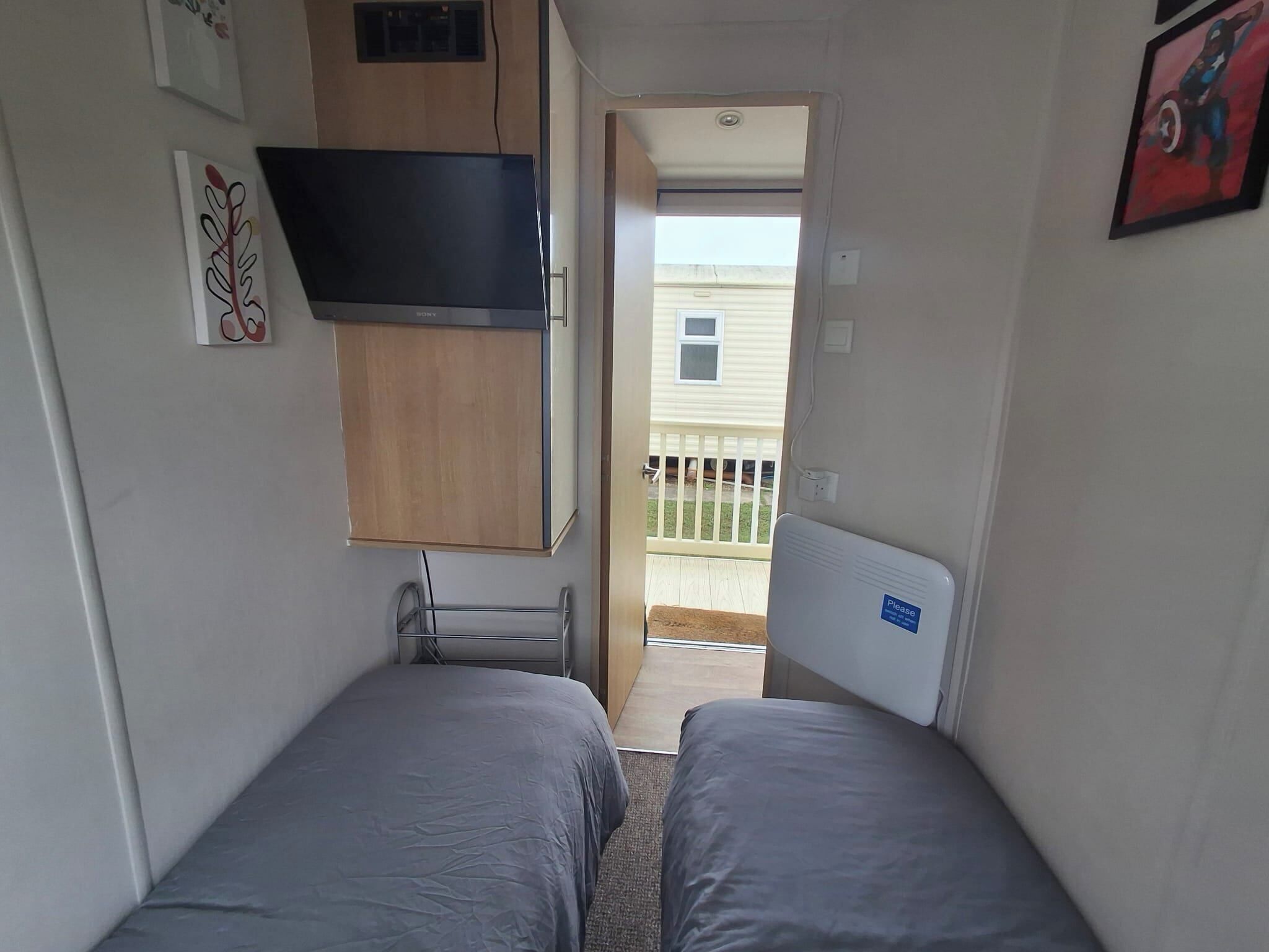 A small, compact room with two single beds, a built-in TV, and a small kitchenette.