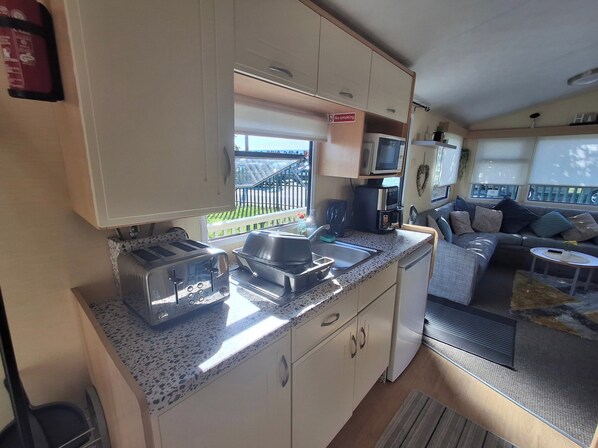A compact kitchen with a microwave, toaster, and sink. There is a small refrigerator and a dining area with a sofa and a coffee table. - 3-bed Caravan - Sleeps 8 - Pool Onsite - Petsok (Skegness)