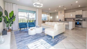 Una sala de estar moderna con un sofá blanco, un sillón azul y una mesa de centro. Hay una zona de cocina con armarios blancos y un microondas.