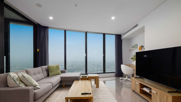 A modern living room with a sectional sofa, a coffee table, a TV on a wooden stand, and large windows offering a city view.