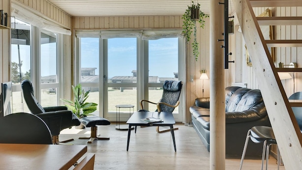 Una habitación amplia con ventanas grandes, techo de madera, sofá de cuero negro, mesa de comedor y una planta colgante.