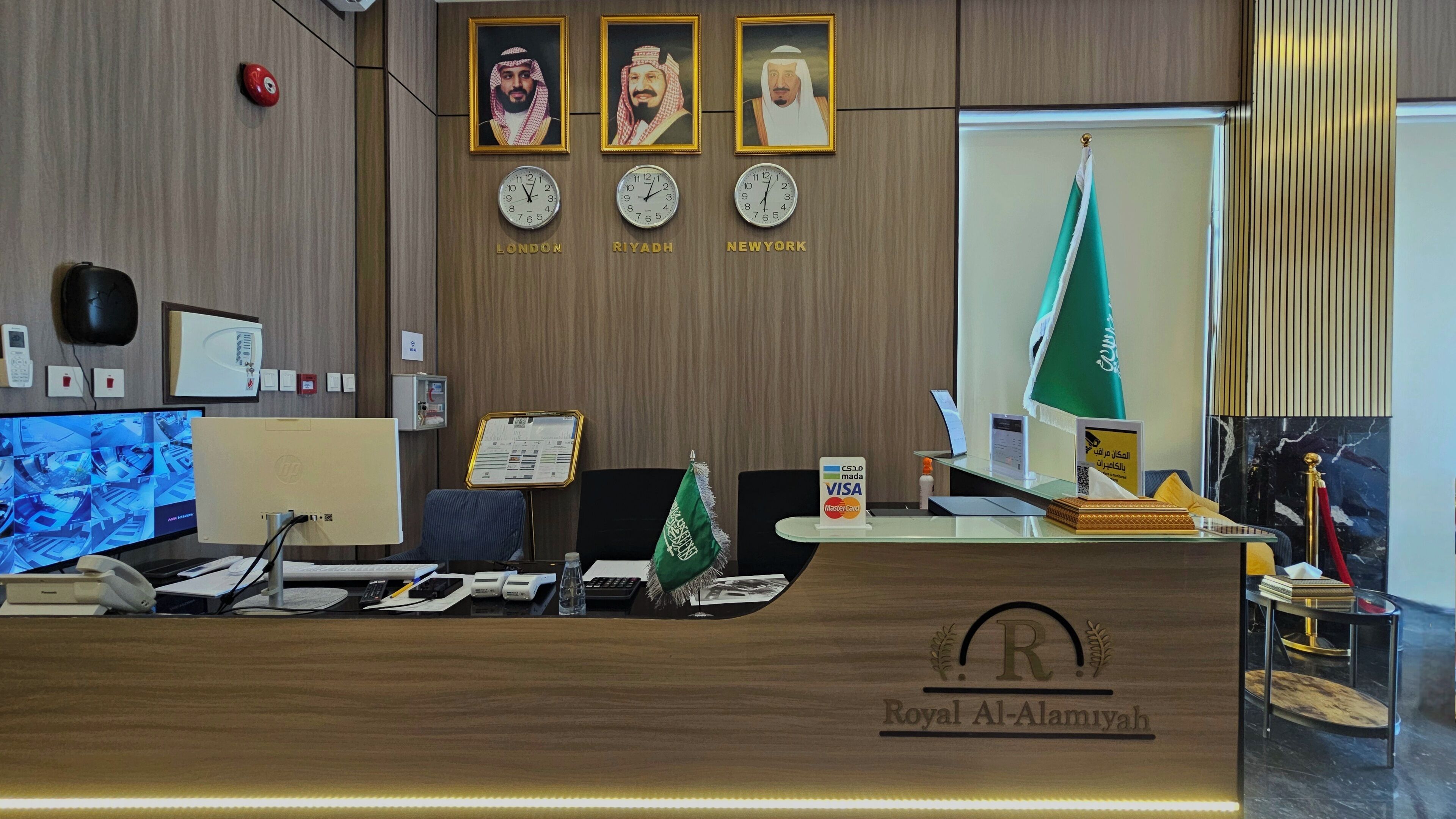 A reception desk with clocks indicating London, Riyadh, and New York times, a flag, and a logo for Royal Al-Aliyah.