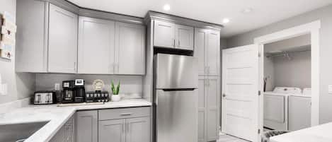 Una cocina moderna con armarios grises, un refrigerador de acero inoxidable, una estación de café y un área de lavandería visible a través de una puerta abierta.