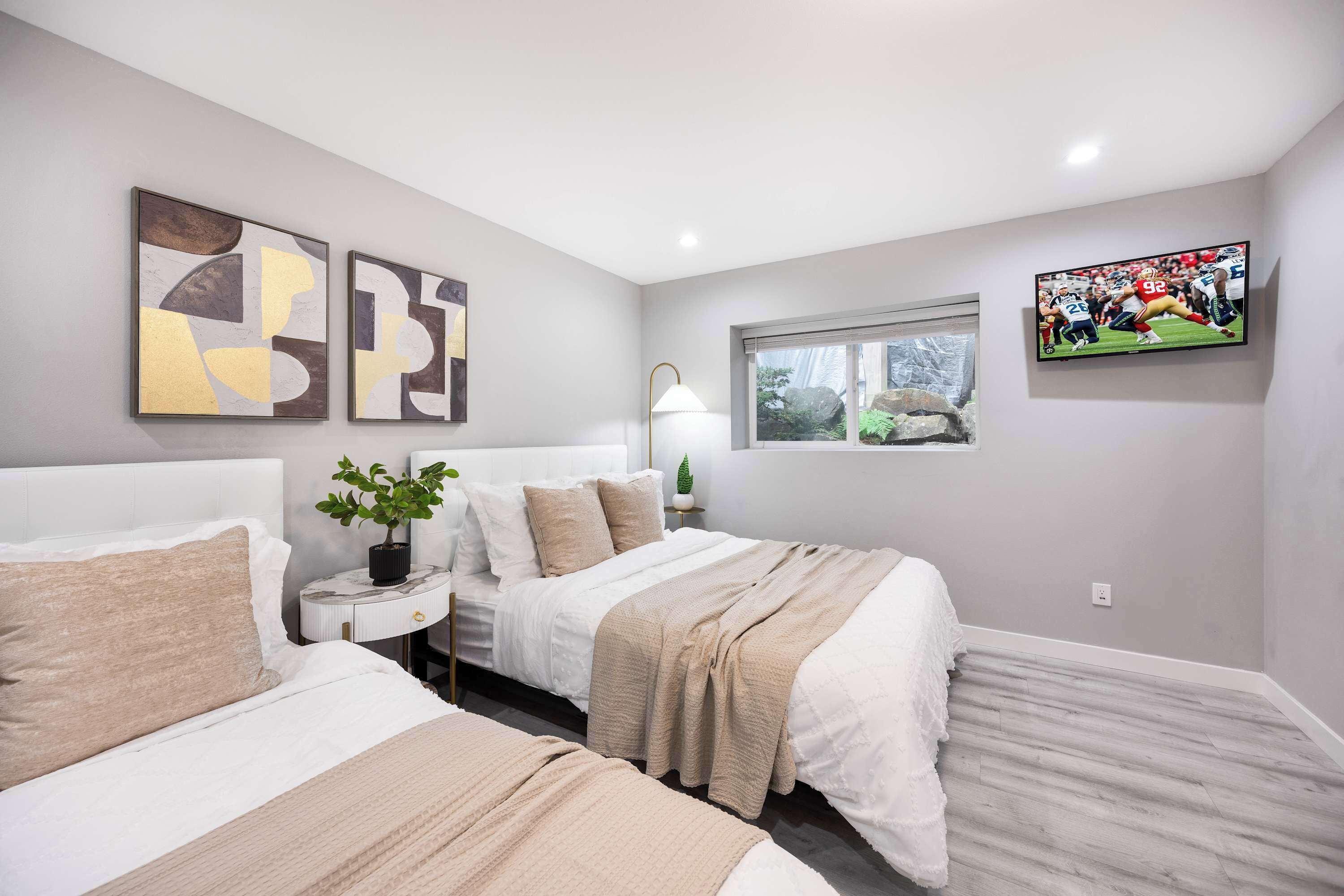 A modern hotel room with two beds, a large window, a flat-screen TV, abstract wall art, and a bedside plant.