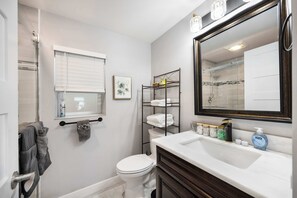 Une salle de bain avec des toilettes blanches, un lavabo avec un miroir, un porte-serviettes et une étagère contenant des articles de toilette.