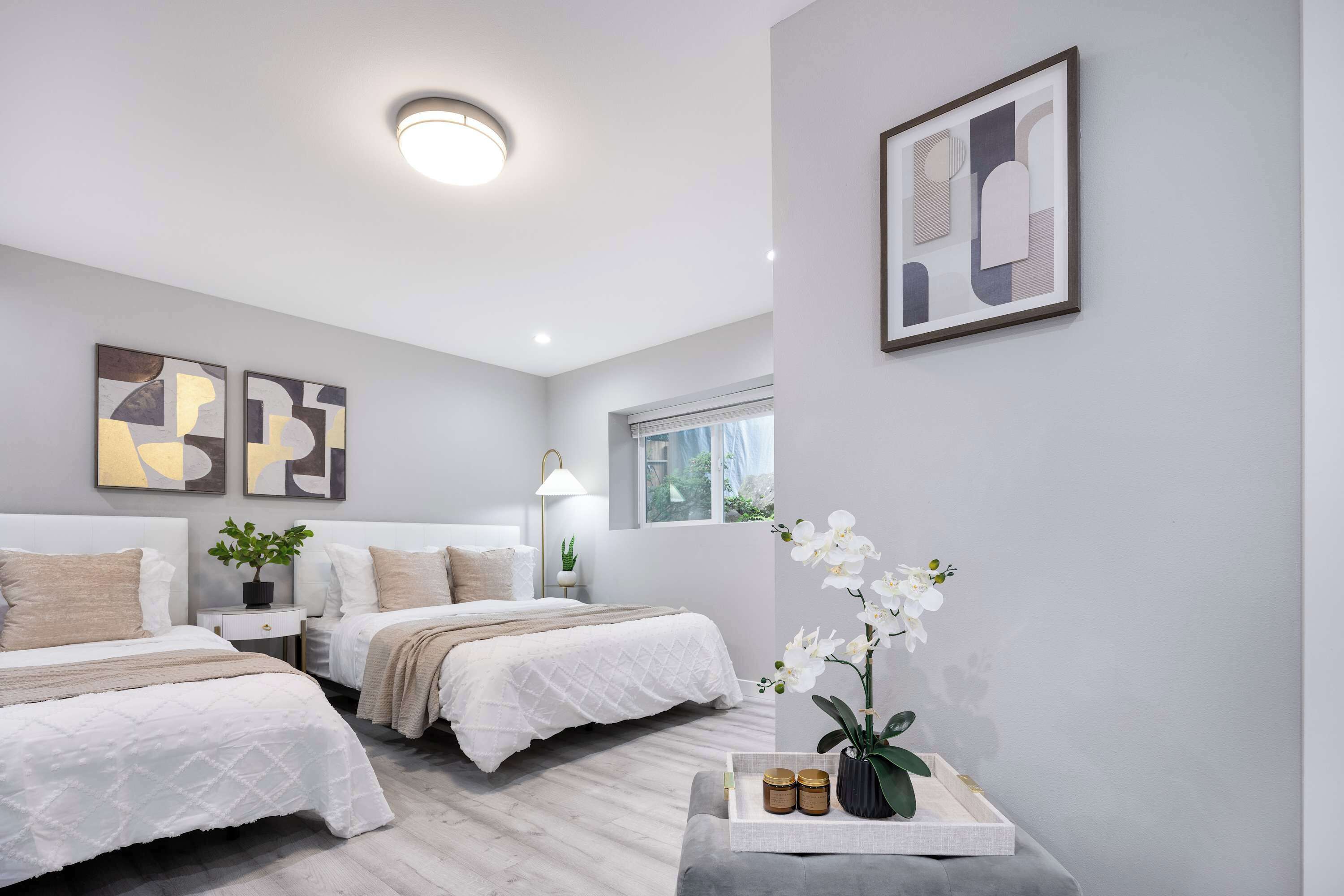 Quarto moderno com duas camas, mesa de cabeceira com abajur, janela com vista e arte abstrata na parede.