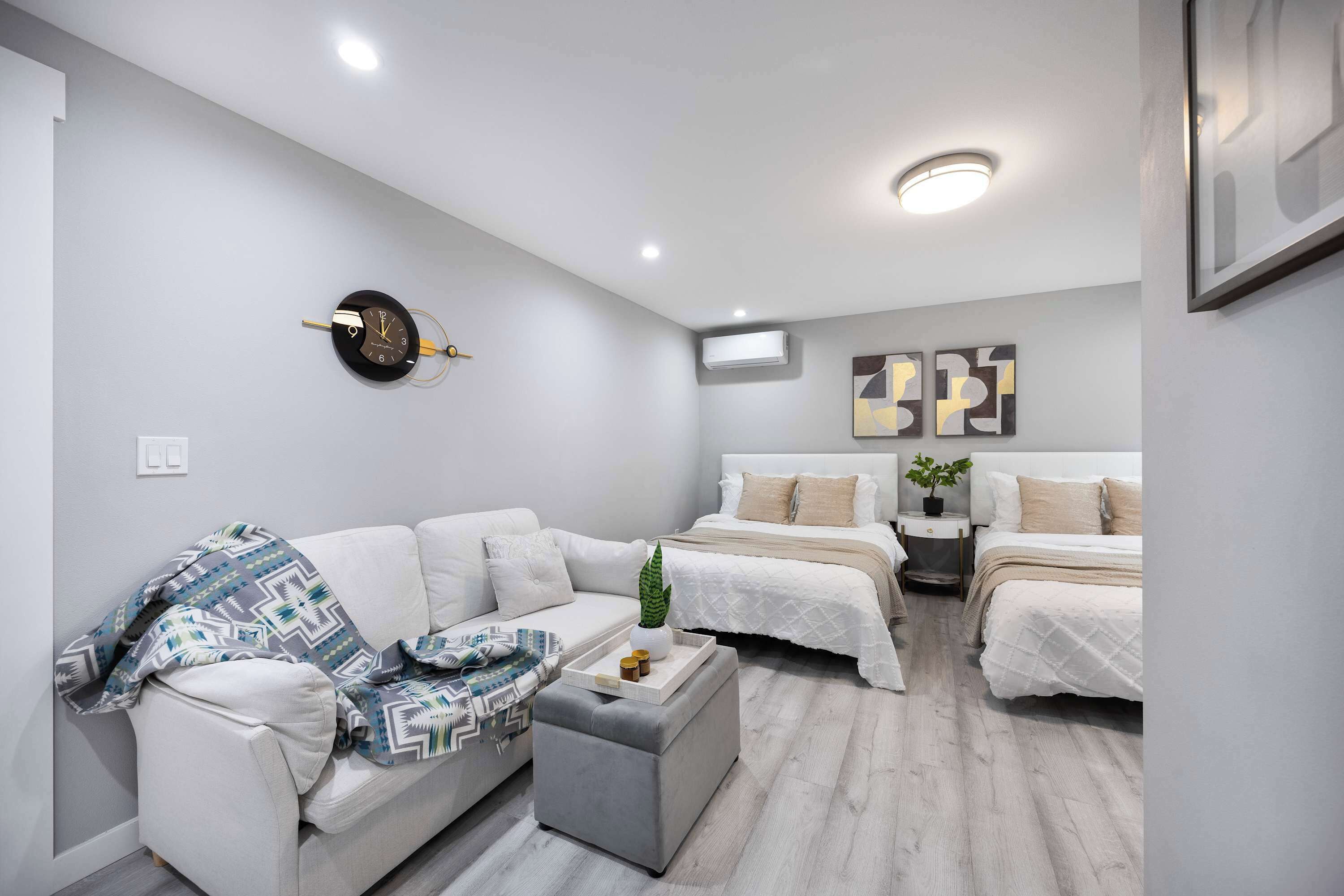 A modern, well-lit bedroom with a grey couch, two single beds, a bedside table with a plant, and a wall clock.