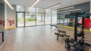 Een ruime fitnessruimte met houten vloeren, krachtapparatuur en grote ramen met uitzicht op palmbomen en gebouwen.