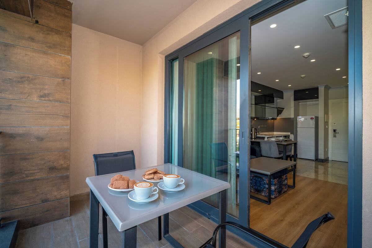 Un área de comedor moderna con un separador de vidrio, una mesa con tres tazas de café y pasteles, y vista hacia la cocina.