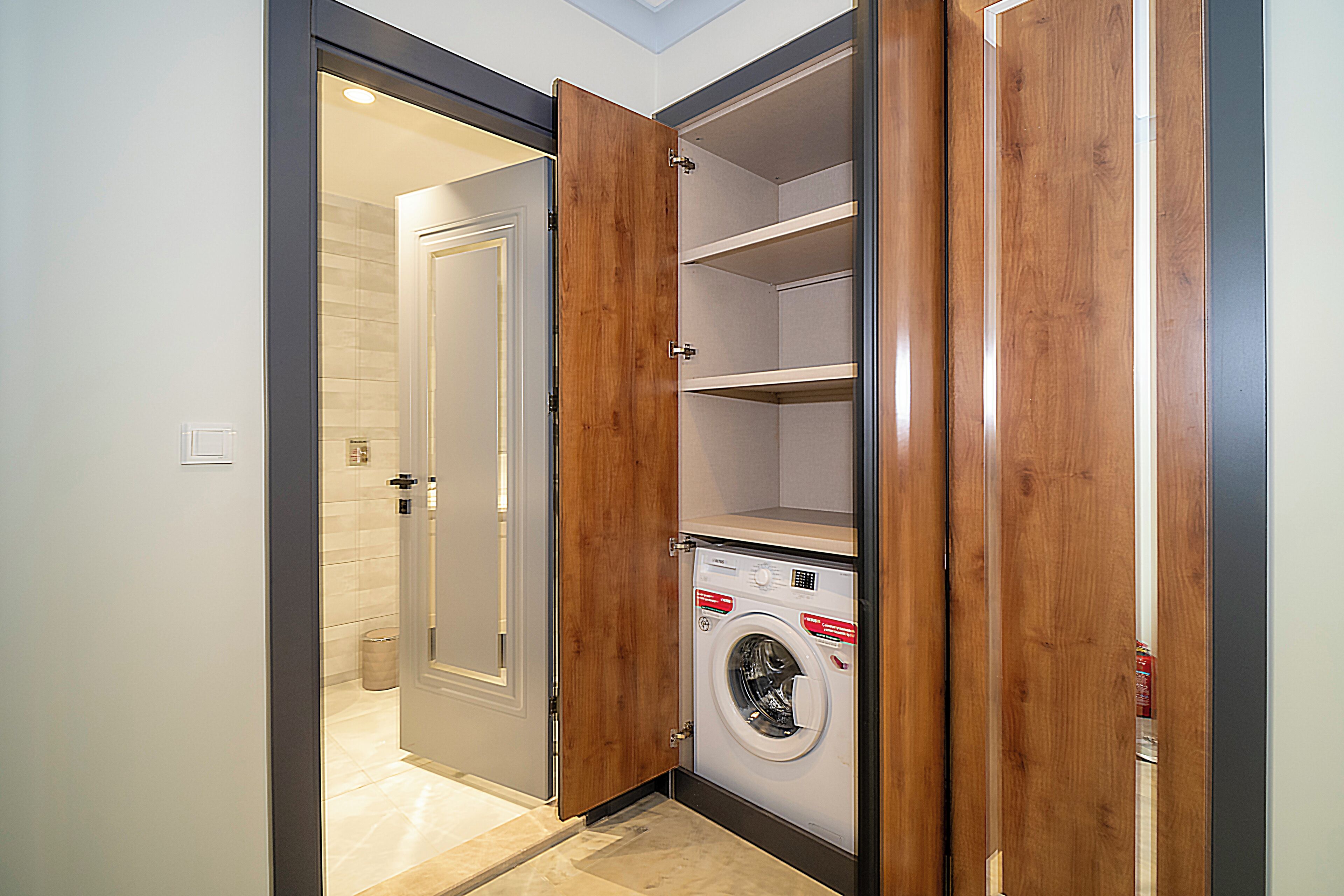 Une petite buanderie équipée d’un lave-linge et d’un sèche-linge encastrés, d’étagères en bois et d’une porte donnant accès à une salle de bain.