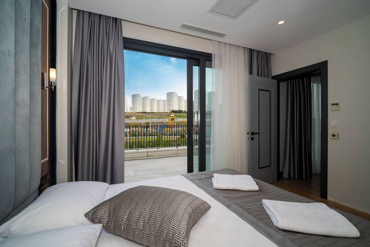 Una habitación de hotel moderna con una cama grande, un balcón con vista a la ciudad y una puerta que da a otra habitación.