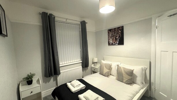 A bedroom with a bed, a bedside table, a lamp, a window with curtains, a radiator, and a leopard print artwork on the wall.
