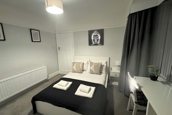 Une chambre d’hôtel avec un lit, une table de chevet et un bureau avec une chaise. Il y a une fenêtre avec des rideaux, un radiateur et un mur orné d’un tableau encadré.