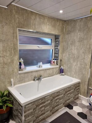 Un baño con pared de textura de piedra, una ventana sobre una bañera y artículos de aseo en el alféizar.