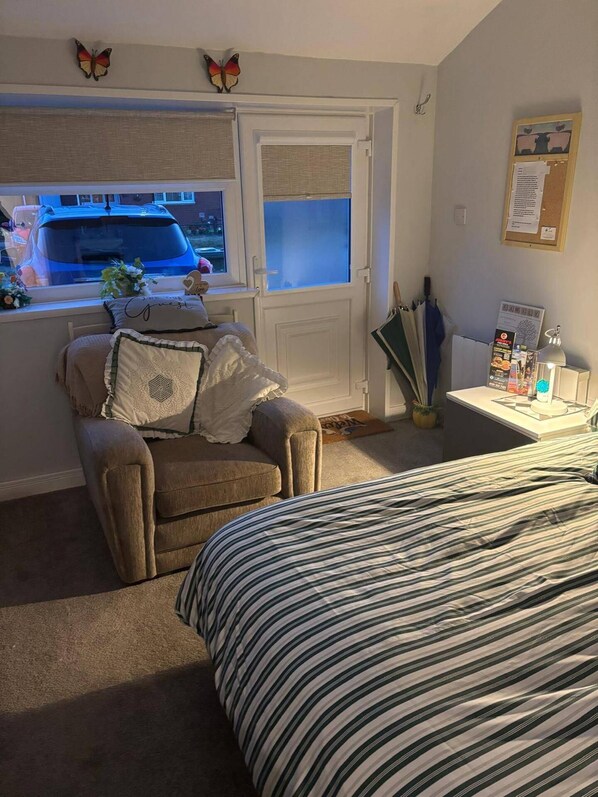 Un dormitorio con cama, una silla, un ventanal y una puerta. Se ve un coche desde la ventana.