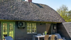 Une maison à toit de chaume avec une porte verte, une table entourée de chaises et un jardin agrémenté de plantes et d’un jardinière en bois.