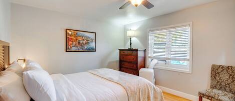 Une chambre à coucher comprenant un lit, un ventilateur de plafond, une fenêtre avec des stores, une chaise et une commode.