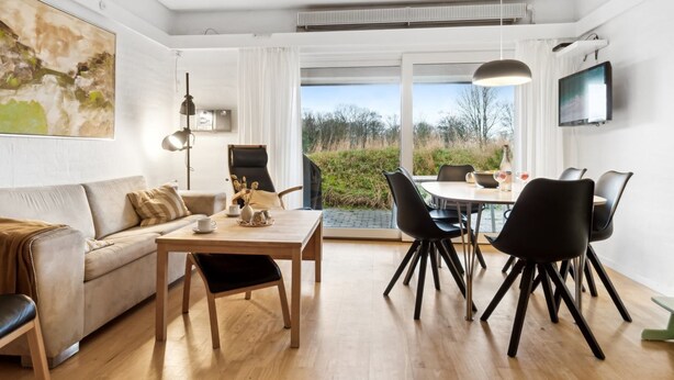 A modern living room with a sofa, a dining table with chairs, a TV mounted on the wall, and a large window offering a view of greenery. - "Answald" - 6km from the sea by Interhome (Aakirkeby)