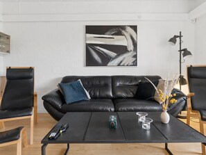 Un salon moderne avec un canapé en cuir noir, deux fauteuils noirs, une table basse noire avec des télécommandes, et une peinture abstraite minimaliste accrochée au mur.