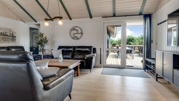 A spacious living area with a leather sofa, a wooden coffee table, and a dining table with chairs. There is a large window with a view of the outdoors, and a sliding glass door leading to a deck.