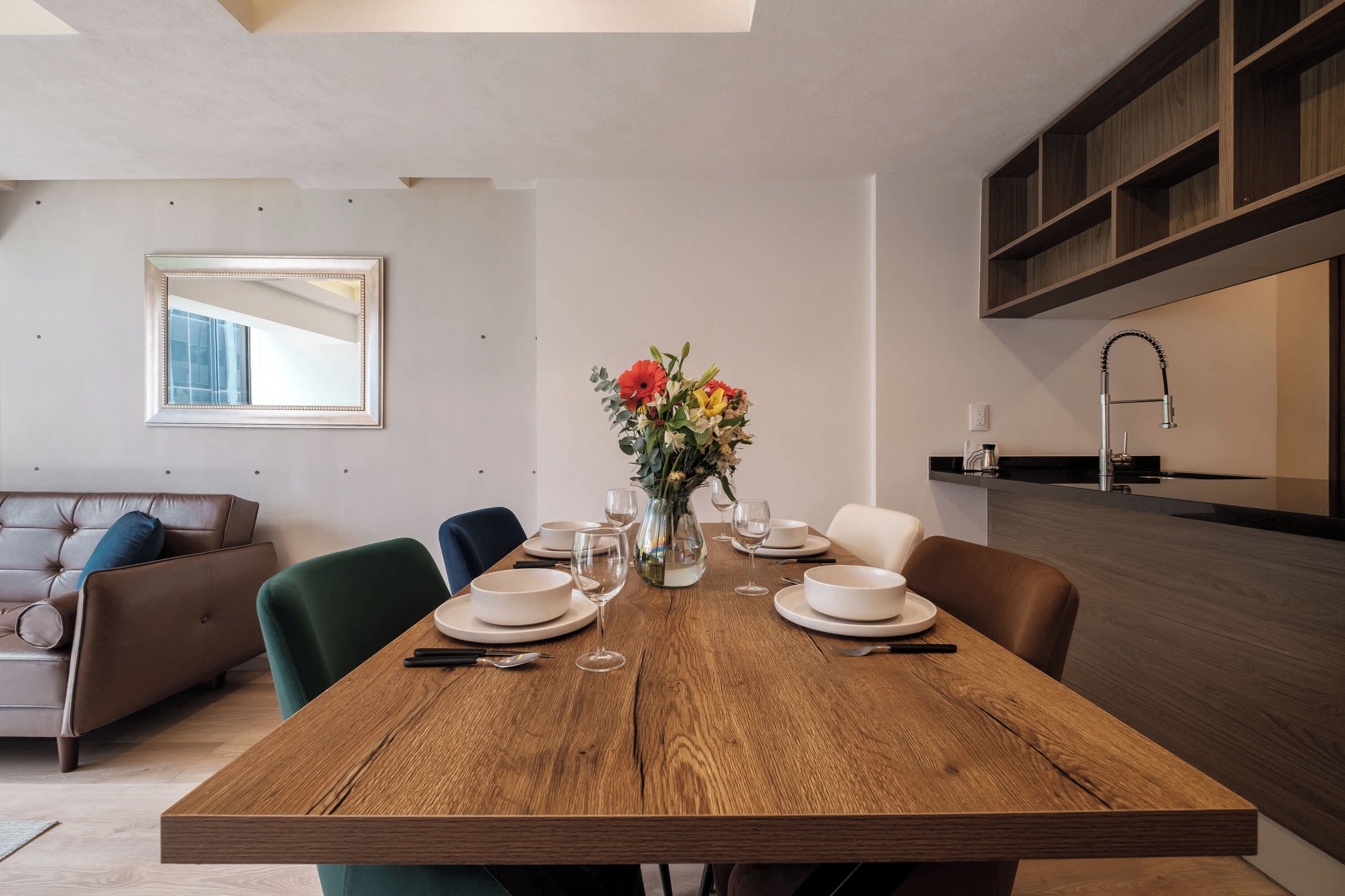 Un comedor con una mesa de madera dispuesta para cuatro, un jarrón de flores y una cocina moderna al fondo.