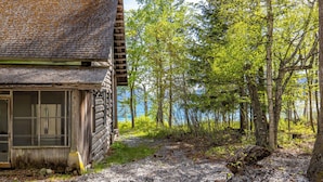 Een landelijke hut met een grindpad er naartoe, omringd door bomen en een wateroppervlak in de verte.