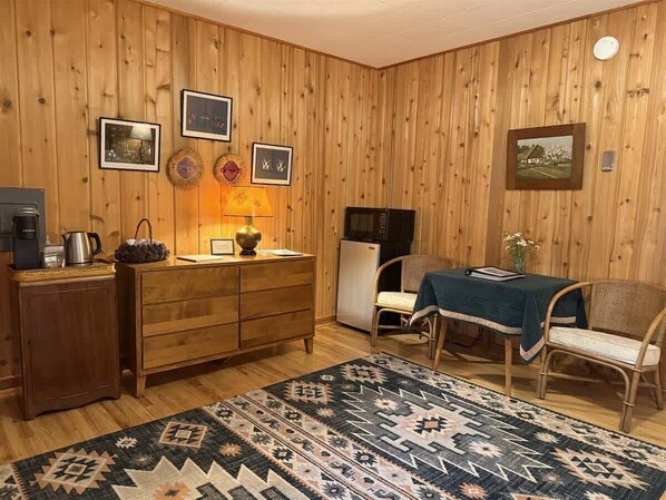 Ein Zimmer mit Holzwandverkleidung, einem hölzernen Schminktisch, einem Esstisch mit einem Stuhl, einem gemusterten Teppich und gerahmten Bildern an der Wand.
