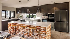 A modern kitchen with dark cabinets, a large island with stools, stainless steel appliances, and pendant lighting. - STYLISH AND SOPHISTICATED TWO STOREY RETREAT WITH SPARKLING POOL (Dunsborough)