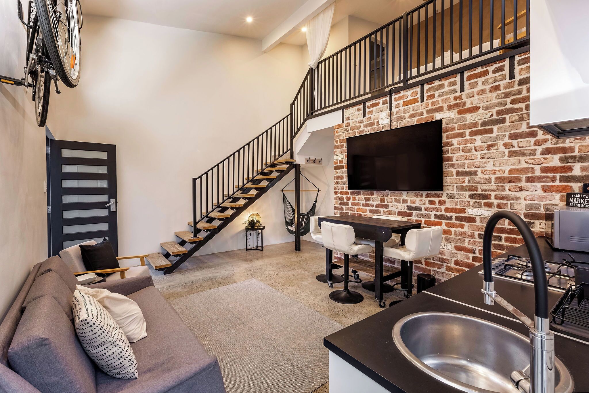 Un espacio habitable moderno con sofá, comedor, cocina con lavaplatos, escalera y pared de ladrillo.