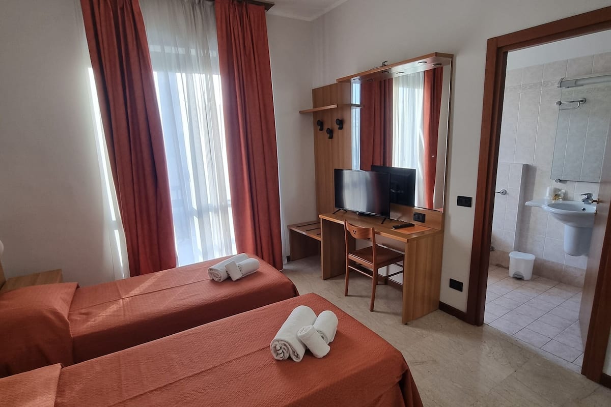 Quarto de hotel com duas camas, uma escrivaninha, televisão e banheiro visível por uma porta aberta.