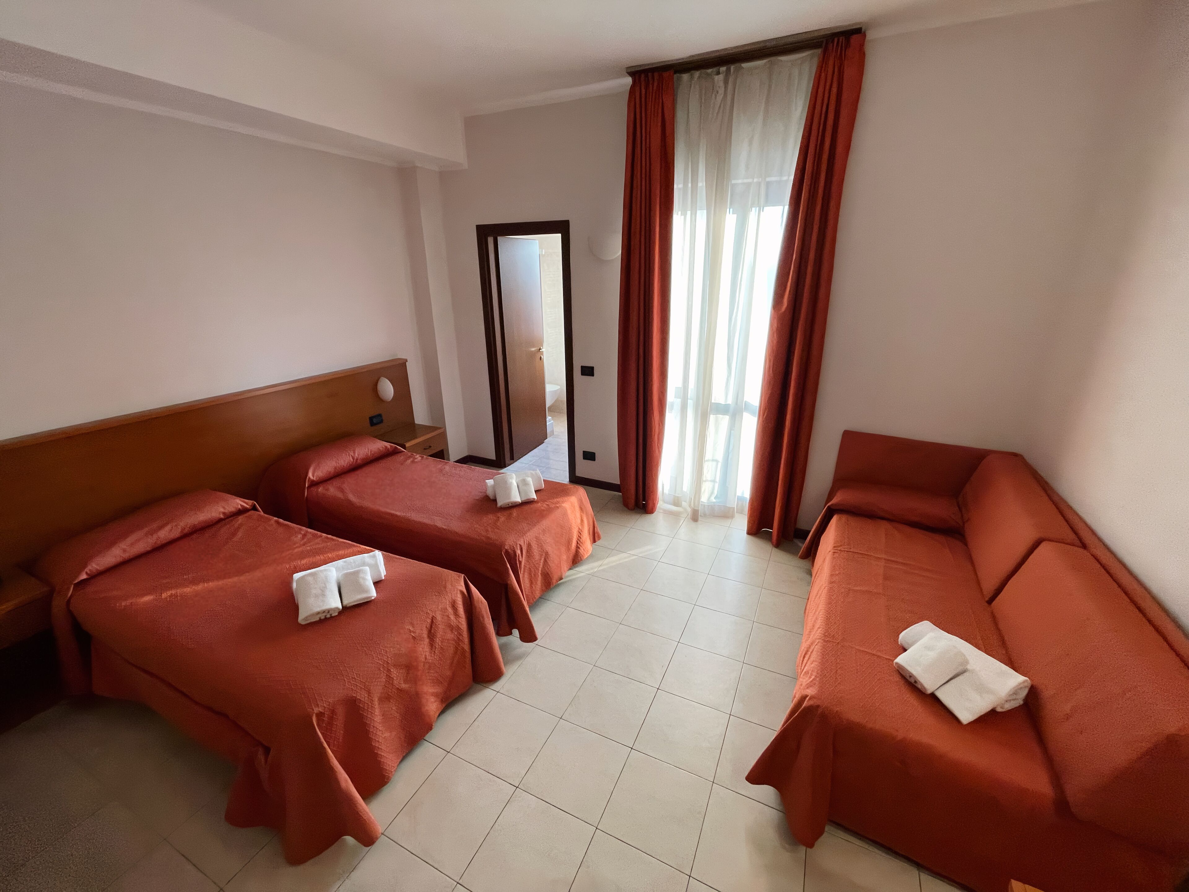 Quarto de hotel com duas camas de solteiro e um sofá-cama, ambos com roupa de cama vermelha e toalhas brancas.