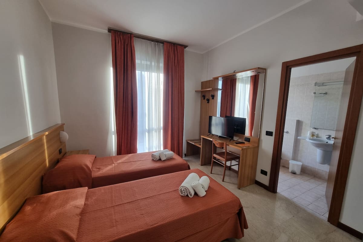 Quarto de hotel com duas camas, uma secretária e televisão, e casa de banho visível através de uma porta aberta.