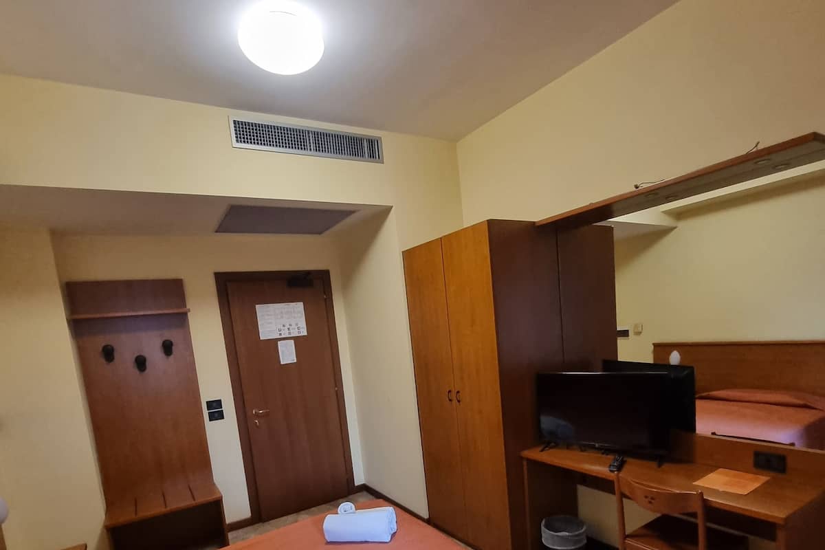 Quarto pequeno e bem iluminado, com cama, escrivaninha, televisão e guarda-roupa.