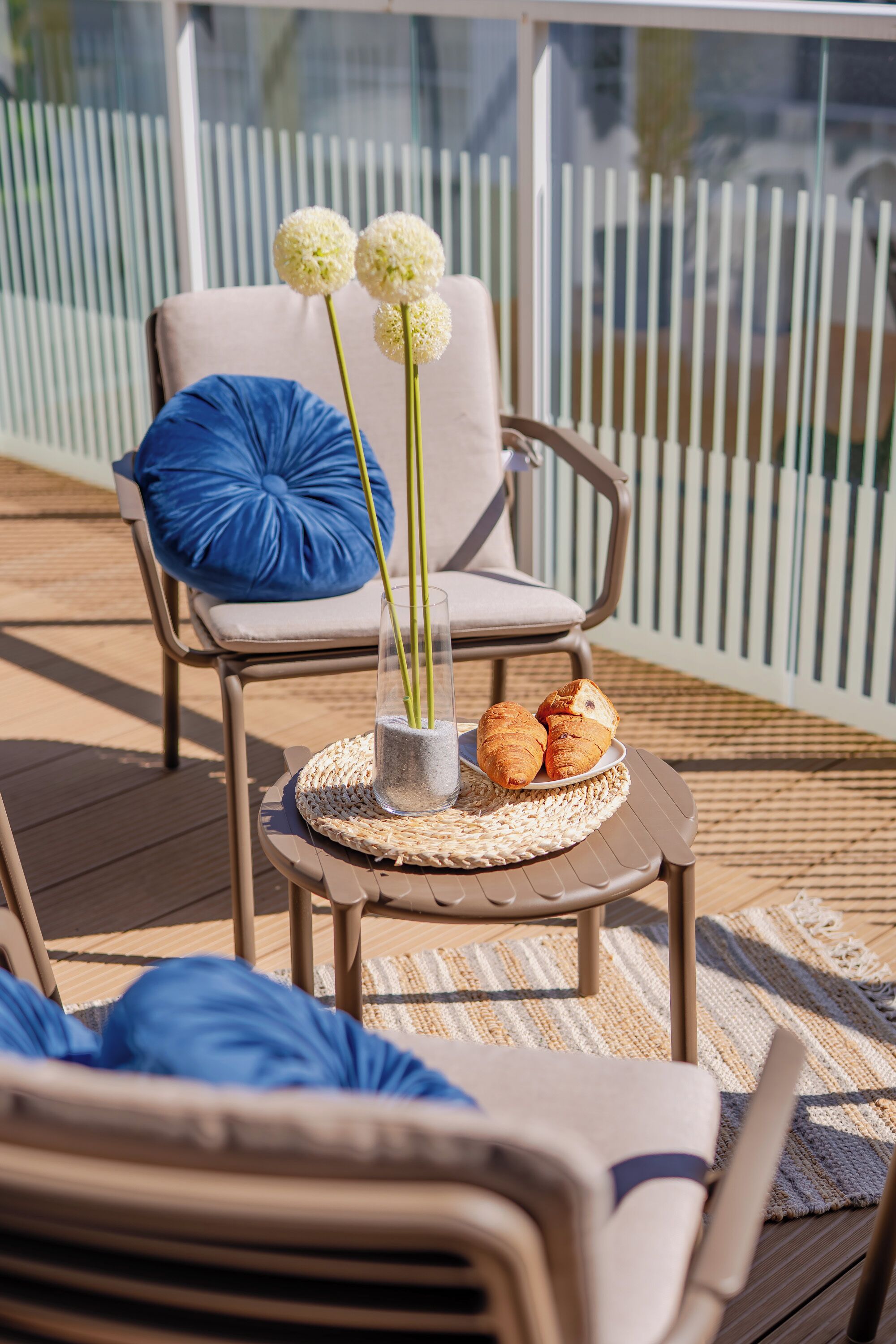 Une terrasse avec une chaise, une petite table ornée d’un vase et de quelques pâtisseries, et un coussin bleu.