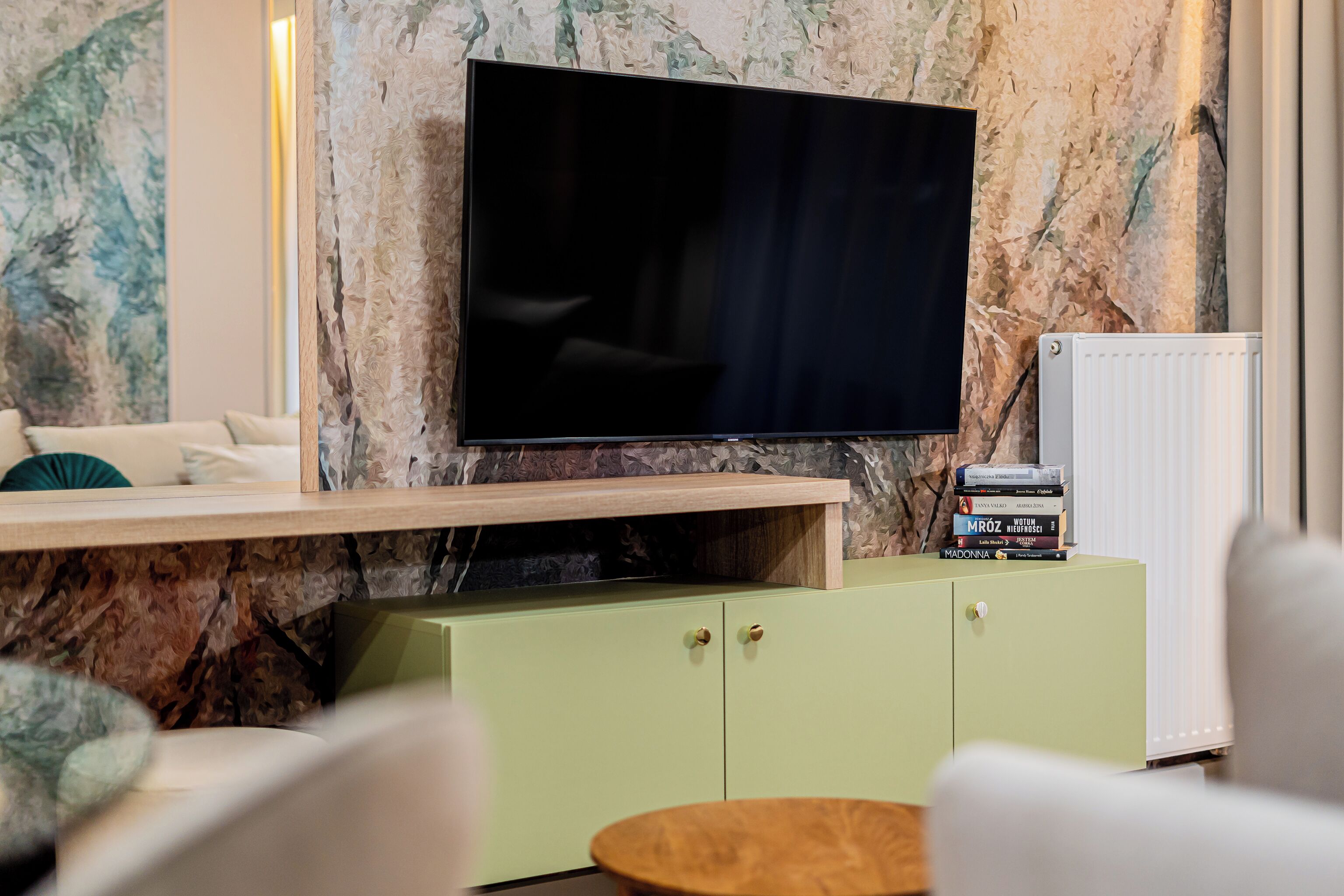 A living room with a flat-screen TV mounted on a marble wall, a green cabinet below, and a round wooden coffee table.