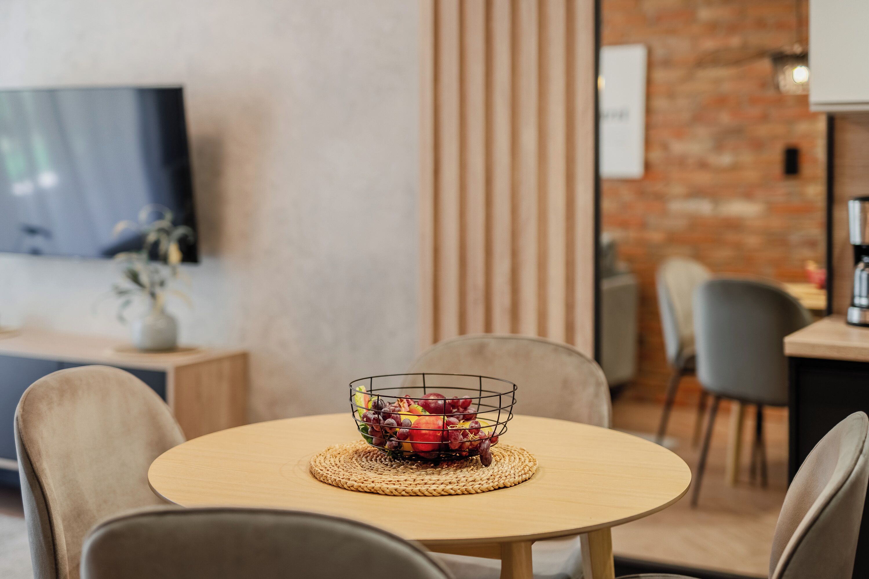 A round wooden table with a woven placemat, surrounded by chairs, in a modern interior with a brick wall and a flat-screen TV mounted on the wall.