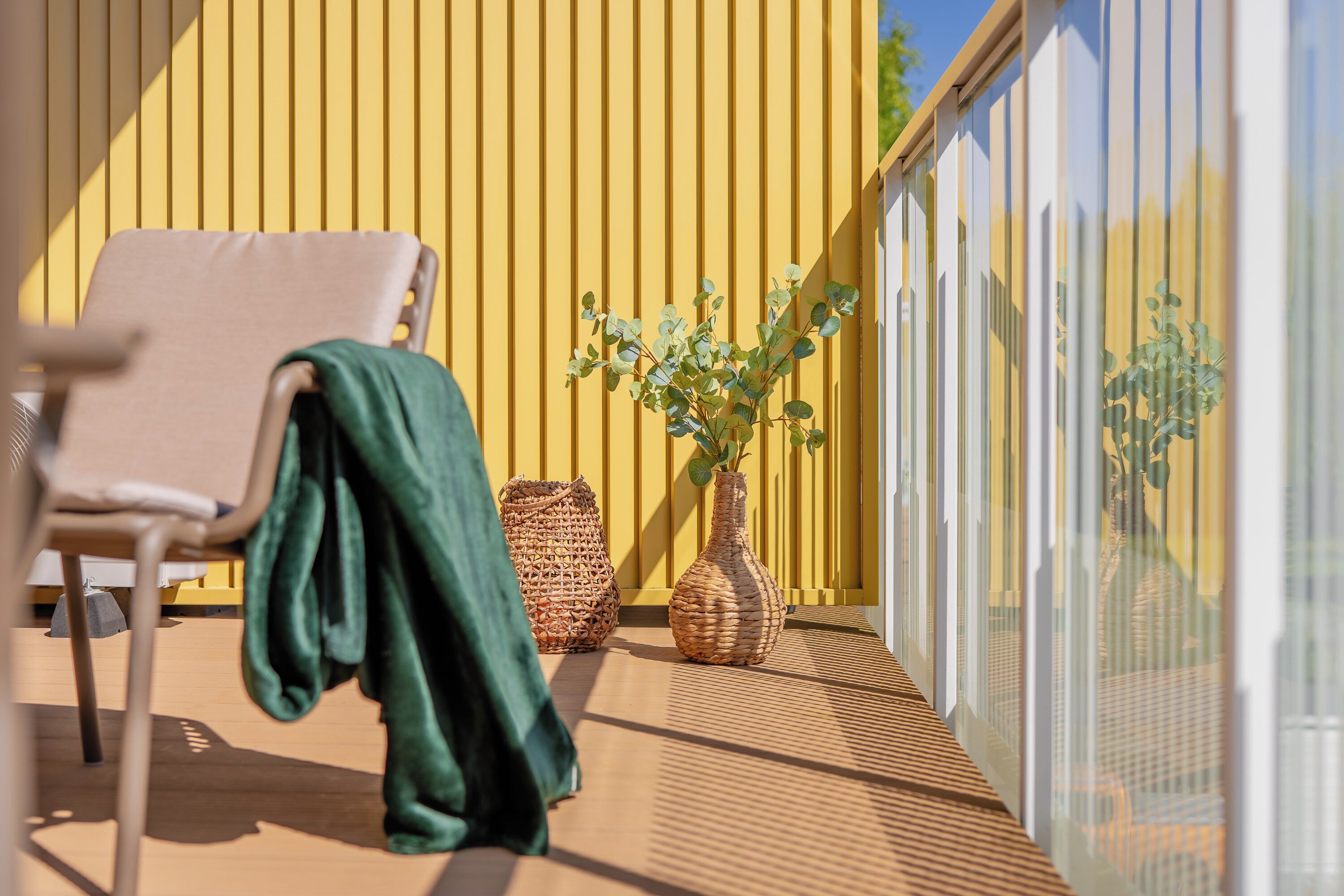 Un balcón con una silla, una toalla verde, una cesta trenzada y un florero de mimbre con plantas.