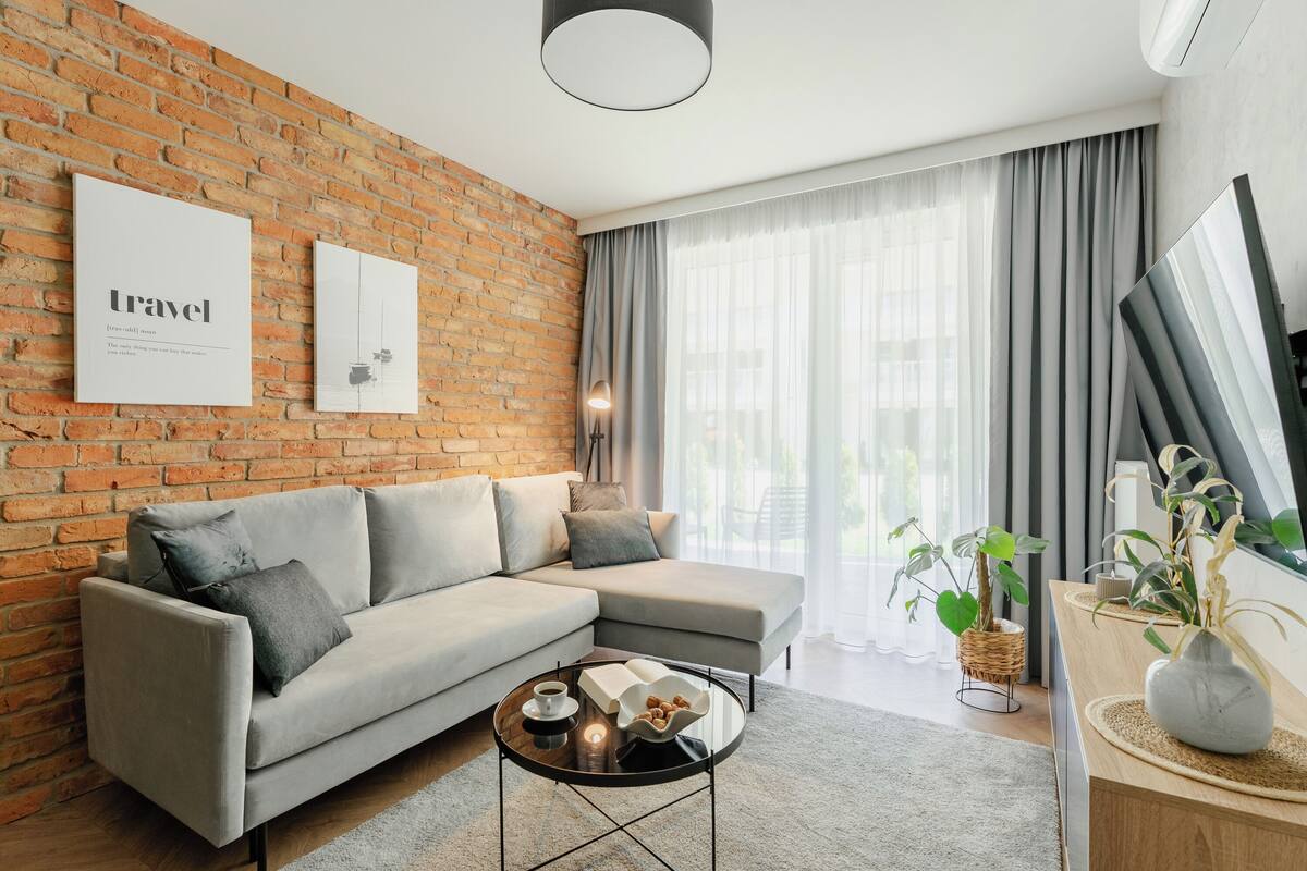 A modern living room with a brick wall, a grey sofa, a coffee table, and a large window with curtains.